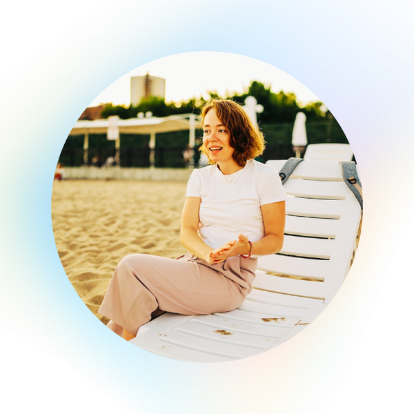 A woman with short brown hair, wearing a white t-shirt and beige pants, sits smiling on a white lounge chair at a sandy beach with greenery and buildings in the background.