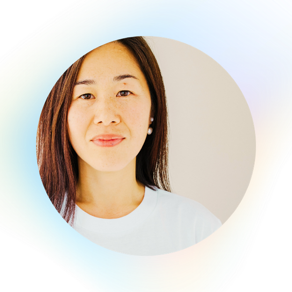 A woman with straight brown hair and a calm expression poses against a light background, partially framed by a faint circular gradient. She wears a white top and pearl earrings.